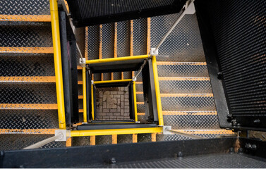 A downward view of a spiral metal staircase with yellow handrails, showing geometric patterns and...