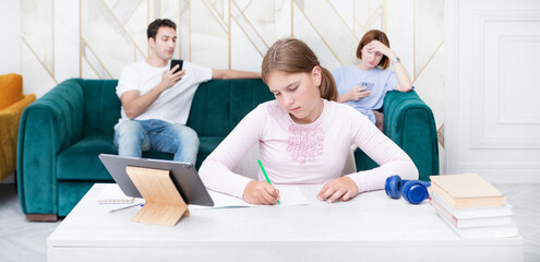 Tired school girl studying hard while her parent having a rest on background	