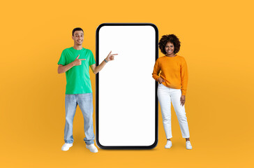 Funny man and woman are standing side by side, both looking at a blank phone screen. The man is...