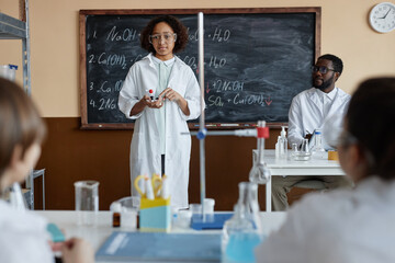 Over-the-shoulder shot African American boy wearing lab coat standing in front of class speaking on subject during Chemistry lesson