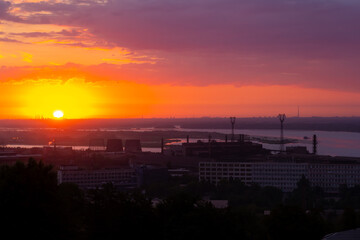 Fototapeta premium Beautiful panoramic cityscape of industrial district during romantic sunrise. Orange sky and clouds. Soft focus. Silhouettes of manufacturing buildings with pipes by river. City landscape theme.