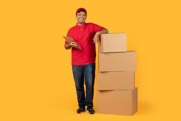 A delivery worker dressed in a red uniform holds a clipboard and leans casually against a stack of...