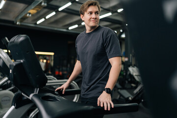 Smiling young man in sportswear walking on treadmill preforming warming-up run routine exercise in...