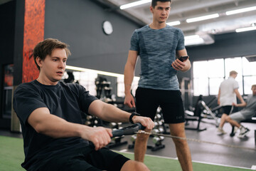 Focused beginner sportsman with personal instructor working out in gym by using rowing machine....