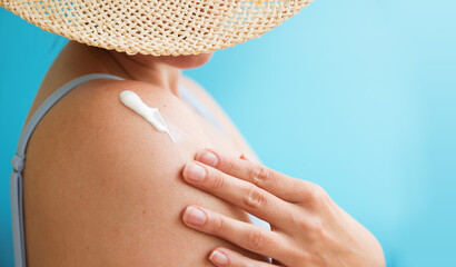 A girl in a straw hat applies sunscreen to her shoulder. Blue background. Summer. Skin protection.