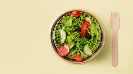 Fresh green salad with tomatoes and cucumbers in a paper container with a wooden fork on a yellow background. Healthy food concept in biodegradable eco-friendly takeaway dishes. Top view.