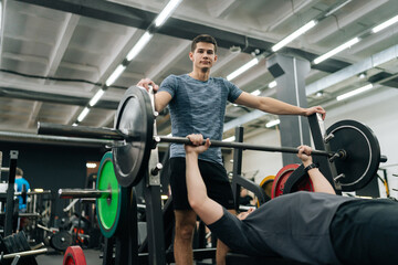Shooting from below of focused beginner male athlete performing incline bench press under watchful eye of personal trainer at gym. Athletic young man pumping up chest muscles during hard training.