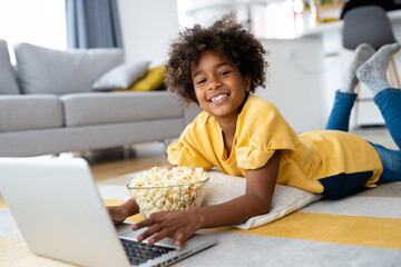 Cheerful African American girl looking at camera while using laptop computer and eating popcorns...