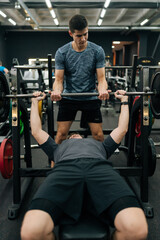 Vertical shot of experienced fitness instructor helping beginner sportsman doing barbell bench...