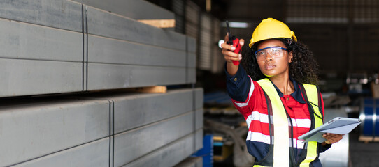 A industrial worker wearing a yellow helmet and a red vest is pointing at something on a clipboard.