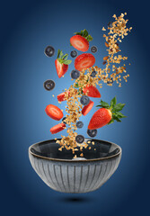 A ceramic bowl in which pieces of strawberries, blueberries, and muesli float in the air and fall,...