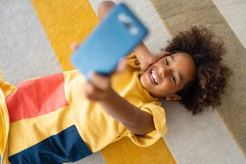 Smiling African American girl using mobile phone while lying on the floor at home.