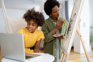 African American mother and daughter painting together at home on a canvas and using laptop for an...