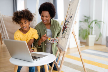 Smiling African American mother and daughter painting together and using laptop in a living room.