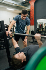 Vertical portrait of personal trainer supporting beginner sportsman in activewear doing bench press with heavy barbell during intense workout in modern fitness center. Concept of healthy lifestyle.