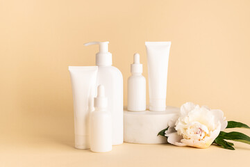 Set of skin care cosmetic products in white plastic unbranded containers on light beige background decorated with aromatic white peony flowers.