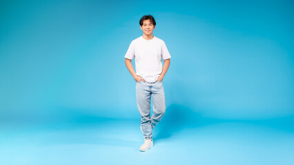 Asian guy in a white t-shirt is standing upright in front of a plain blue background. His posture...