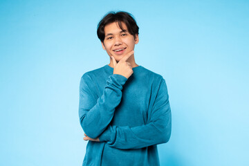 Asian young guy wearing a casual blue sweater places one hand on his chin, striking a thoughtful...