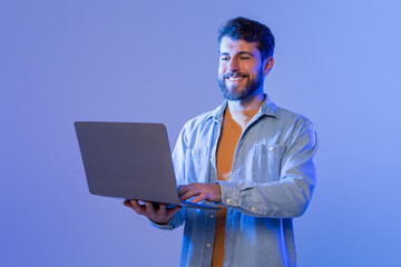 A man sitting at a desk is smiling while interacting with a laptop computer. He appears engaged and focused on the screen, possibly working or communicating with others online.