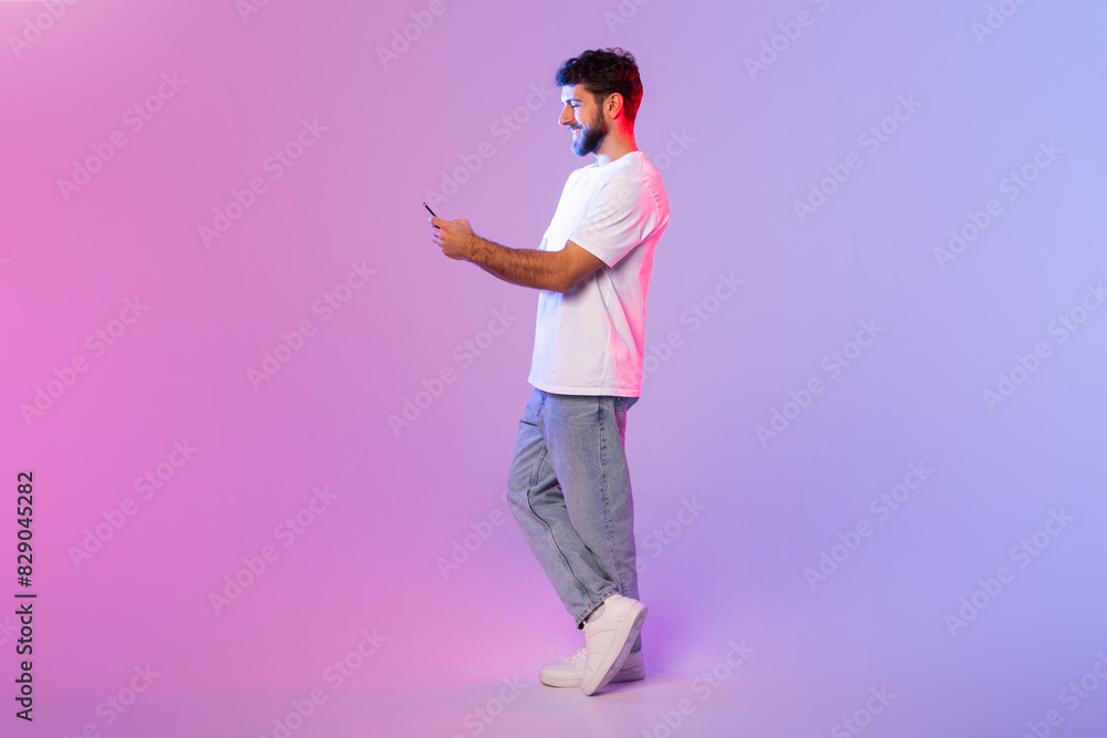Poster A man dressed in a white t-shirt is holding a cell phone in his hand, looking at the screen. He appears focused on whatever he is viewing or doing on the device.