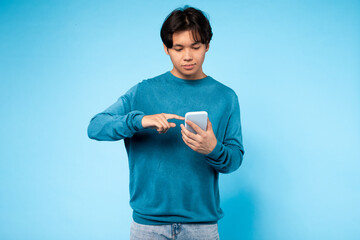 Asian guy wearing a blue sweater is focused on his cell phone screen, tapping and scrolling with...