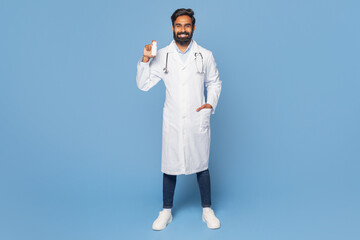 A bearded Indian doctor wearing a white lab coat and stethoscope smiles confidently while holding a...