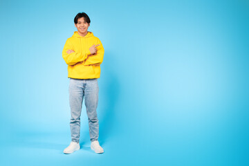 Fototapeta premium Asian guy wearing a yellow hoodie is standing with his arms crossed, pointing at copy space. He appears confident and assertive.