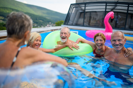 Group of cheerful seniors shaving fun in pool jumping, swiming and lounging on floats. Elderly friends spending hot day by swimming pool.