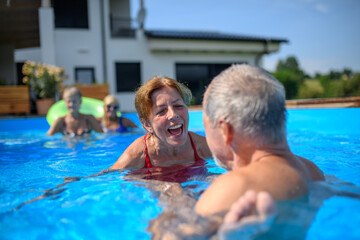 Couple of cheerful seniors shaving fun in pool with friends jumping, swiming and lounging on floats. Elderly friends spending hot day by swimming pool.