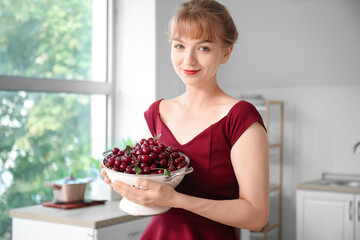 Happy beautiful young woman with colander full of ripe cherries in kitchen