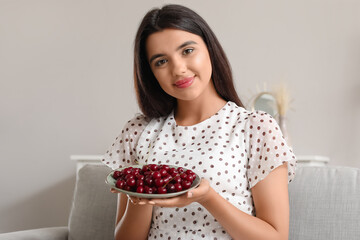 Happy beautiful young woman with bowl of ripe cherries sitting on sofa in living room