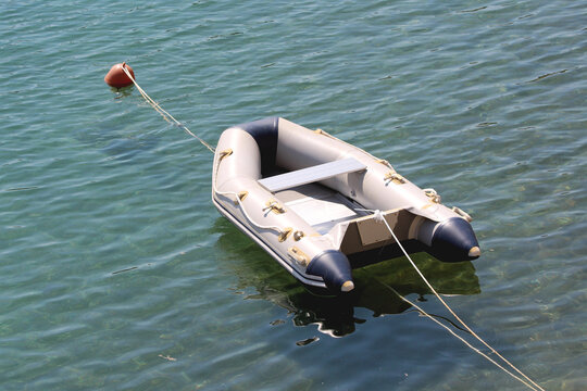Small inflatable grey and dark blue rubber flatbeat dinghy boat with high quality reinforced aluminum floor tied to orange navigational buoy and anchored in local harbor surrounded with calm blue sea 