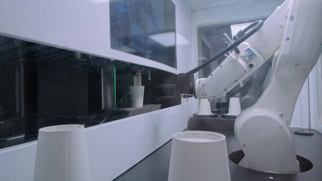 White pick and place robotic arm manipulator with clamp is preparing coffee and moving plastic coffee cups at robot exhibition, trade show: close up. Futuristic and automated technology concept