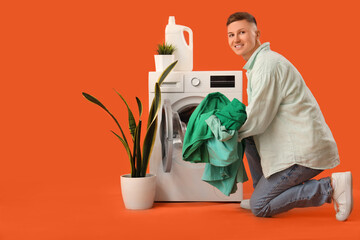 Handsome young man putting laundry into washing machine on orange background