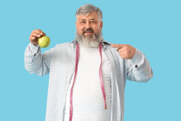Overweight mature happy man with measuring tape pointing at apple on blue background. Weight loss...