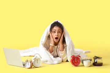 Overslept young African-American woman with blanket, breakfast, laptop and alarm clocks on yellow...