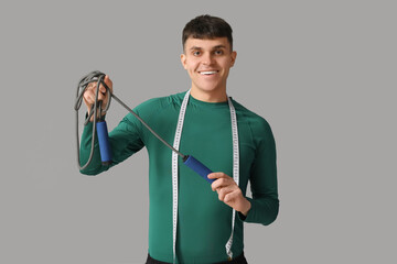Sporty young man with measuring tape and jumping rope on grey background. Weight loss concept