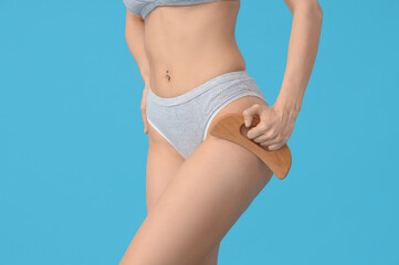 Young woman in underwear massaging her thigh with wooden scraper on blue background, closeup