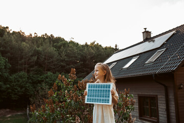 Little girl with model of solar panel, standing in the middle of meadow, house with solar panels...