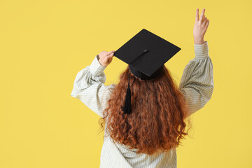 Female student in graduation hat showing victory gesture on yellow background , back view