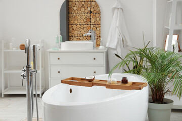 Wooden tray with coconut and cosmetic products on white bathtub in modern bathroom