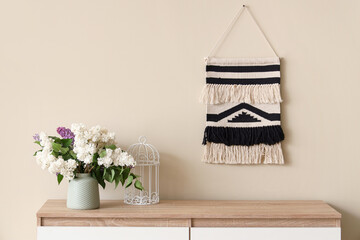 Vase with lilacs on modern chest of drawers near beige wall in room, closeup