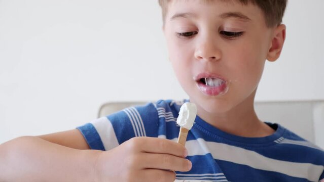 Handsome boy eating popsicle on a stick. He bites off the ice cream with pleasure. Close-up. Child eat in the kitchen or at the dining room table. Tasty dessert. Slow motion. High quality 4k footage