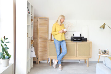 Mature woman with cup of coffee and printer on commode at home