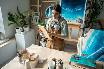 Interested woman painter holding wooden board in hands preparing for epoxy resin application...