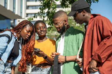 Interested stylish group of african young people students looks at smartphone while walking in...