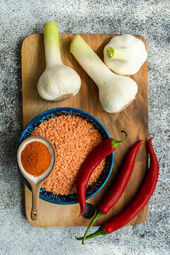 Overhead view of a bowl of red lentils, red chilli peppers, chilli powder and garlic on a wooden chopping board