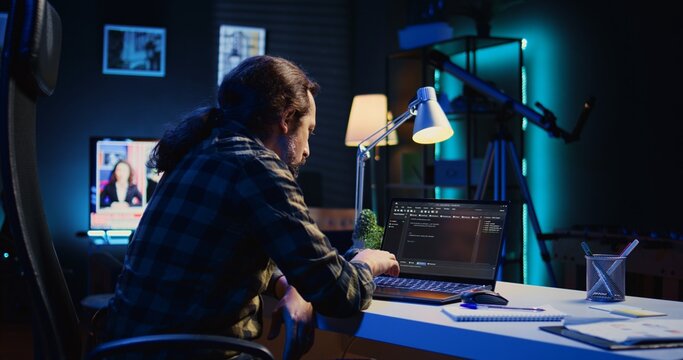 Experienced programmer writes code on laptop screen while in apartment office using Java programming languages. Developer remotely working on fixing database errors while working from home, camera B