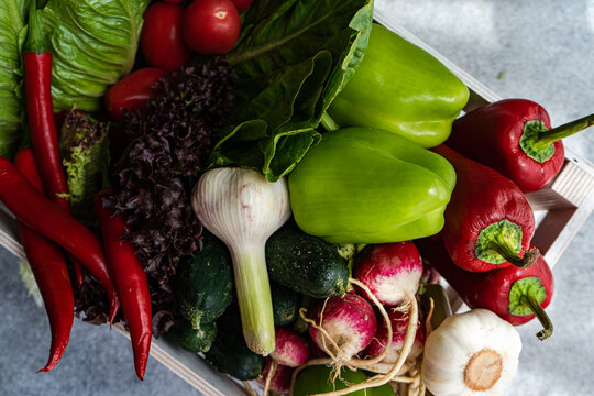 Overhead view of a wooden crate filled with fresh lettuce, bell peppers, chillis, tomatoes, radishes, cucumber and garlic