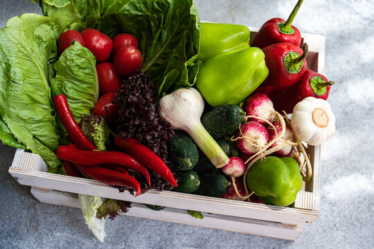 Overhead view of a wooden crate filled with fresh lettuce, bell peppers, chillis, tomatoes, radishes, cucumber and garlic